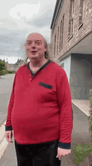 a man with long hair is standing in front of a building wearing a red shirt .