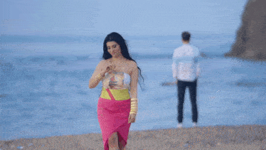 a woman in a pink skirt stands on a beach while a man stands behind her
