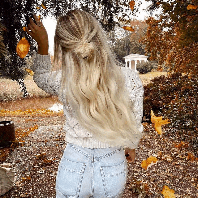 a woman wearing a white sweater and blue jeans is standing in front of a tree with leaves falling from it