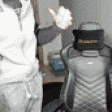 a man in a white jacket is standing in front of a chair with his hand up .