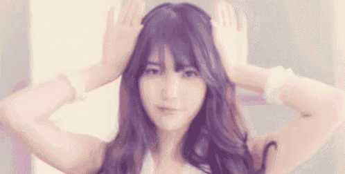 a young woman with long purple hair is making a funny face with her hands on her head .