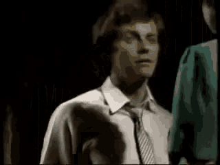 a man in a white shirt and tie is standing next to another man in a green shirt in a dark room .