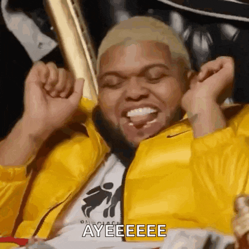 a man in a yellow jacket is sitting in a chair with his hands in the air and the words ayeeeee on his face .