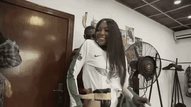 a woman in a white shirt is standing in a room with a fan .