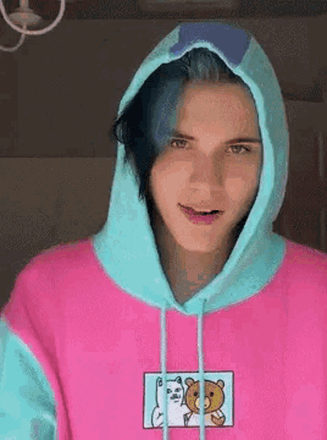 a young man with blue hair is wearing a pink and blue hoodie and smiling .