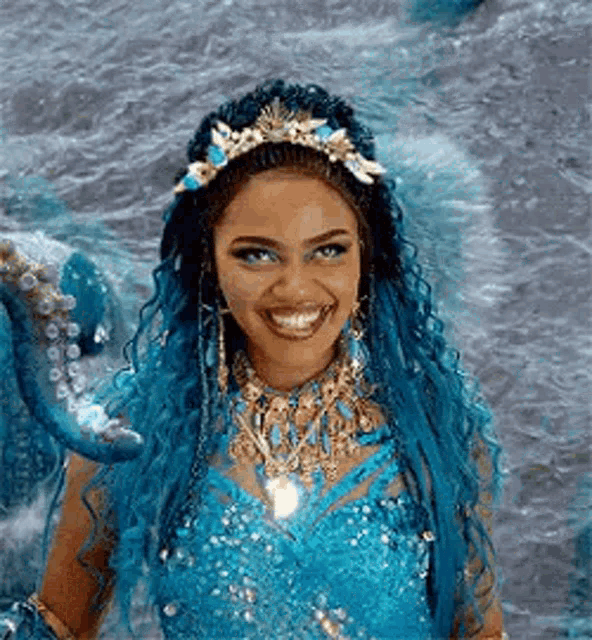 a woman with blue hair and a crown on her head is smiling in front of a waterfall .