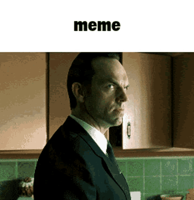 a man in a suit and tie is standing in a kitchen with a meme written above him