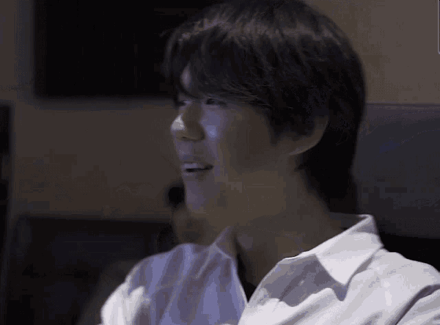 a man in a white shirt is sitting in a dark room and smiling