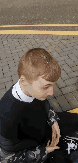 a young man sitting on a brick sidewalk wearing a black sweater with a white collar