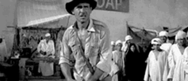 a black and white photo of a man in a hat standing in front of a crowd of people .