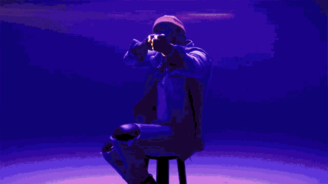 a man is sitting on a stool in front of a purple background and pointing at the camera