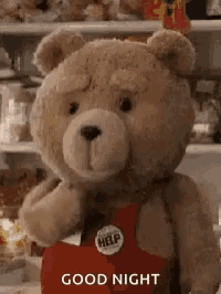 a teddy bear wearing a red apron and a help button is standing in front of a shelf .