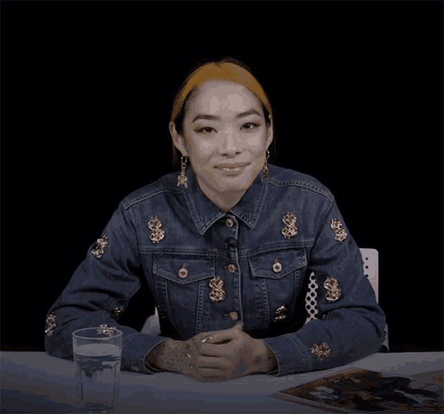 a woman in a denim jacket with dollar signs on it sits at a table with a glass of water