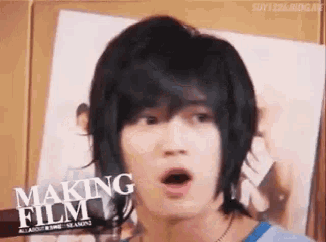 a young man with long black hair is making a funny face while standing in front of a poster .