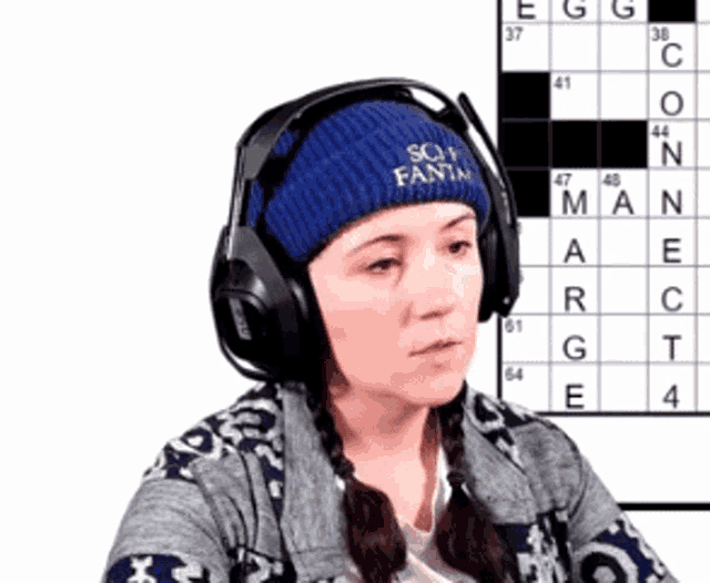 a woman wearing headphones and a blue hat that says soy fanta is sitting in front of a crossword puzzle