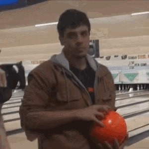 a man in a brown jacket is holding an orange bowling ball