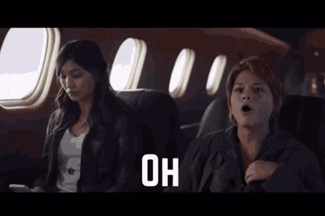 two women are sitting on an airplane and the word wait is on the screen behind them