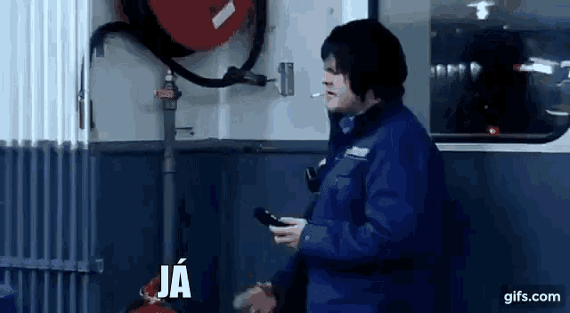 a man in a blue jacket is smoking a cigarette while holding a cell phone in a garage .