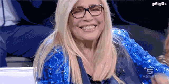 a woman wearing glasses and a blue sequined jacket is smiling on a screen that says mediaset