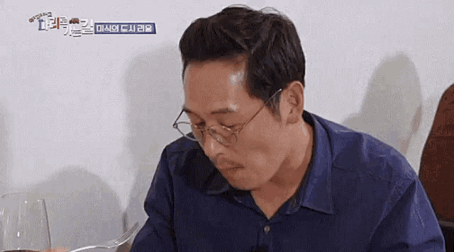 a man wearing glasses and a blue shirt is sitting at a table with a glass of wine