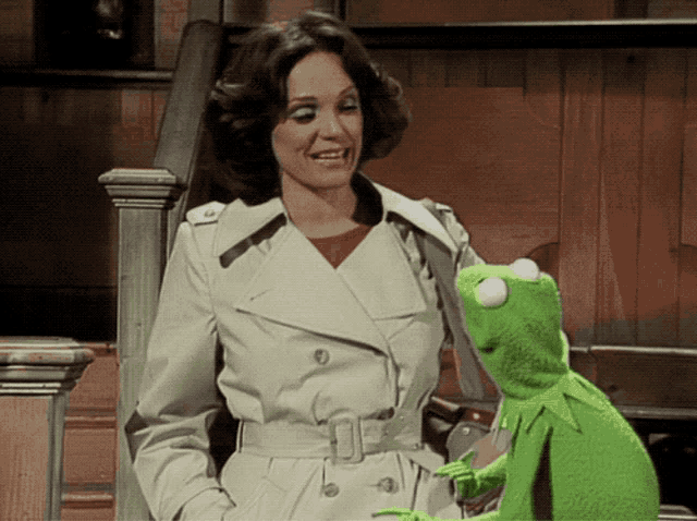 a woman in a trench coat is standing next to a green kermit the frog