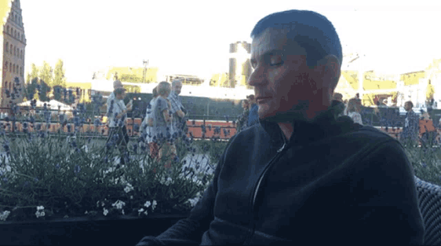 a man in a black jacket sits in front of a planter of purple flowers