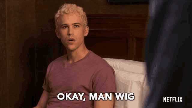 a man is sitting on a bed with okay man wig written on his shirt