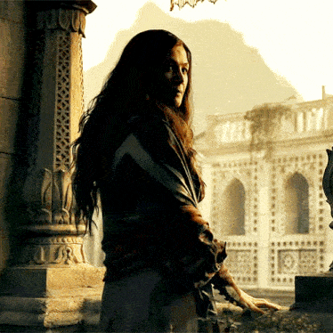 a woman with long hair is standing in front of a building with a mountain in the background
