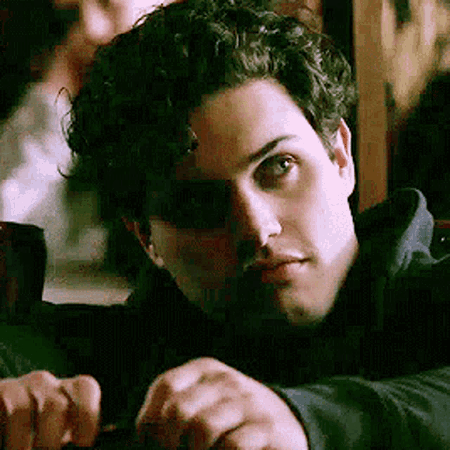 a man with curly hair is sitting at a table and looking at the camera with his hand on his chin .