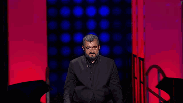 a man with a beard in a black jacket stands in front of a red and blue background