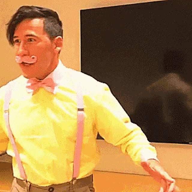 a man wearing a yellow shirt and pink suspenders is standing in front of a tv