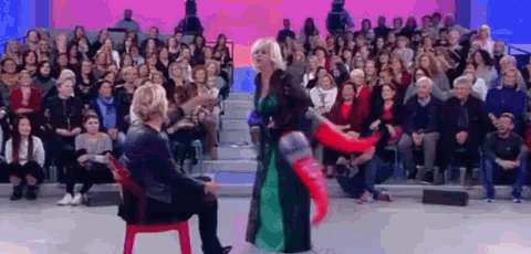 a woman in a green dress is standing next to a woman in a red chair in front of a crowd of people .