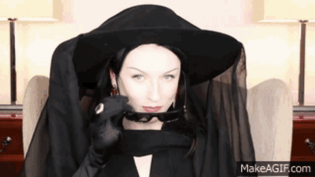a woman wearing a black hat and gloves is sitting in a chair and holding a pair of sunglasses in her mouth