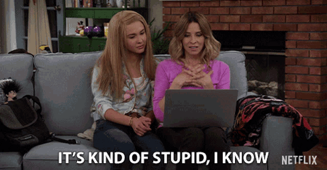 two women sit on a couch looking at a laptop and one of them says it 's kind of stupid