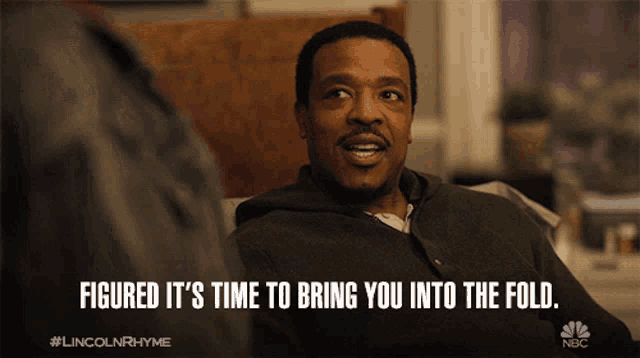 a man is sitting on a couch with the words " figured it 's time to bring you into the fold "