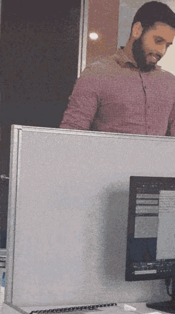 a man in a pink shirt sits at a desk in front of a computer monitor