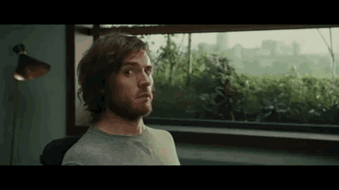 a man with a beard is sitting in front of a window looking out .