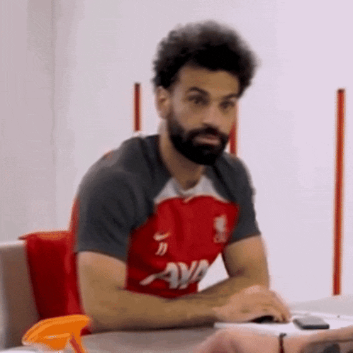a man with a beard is sitting at a table wearing a red shirt that says ava on it