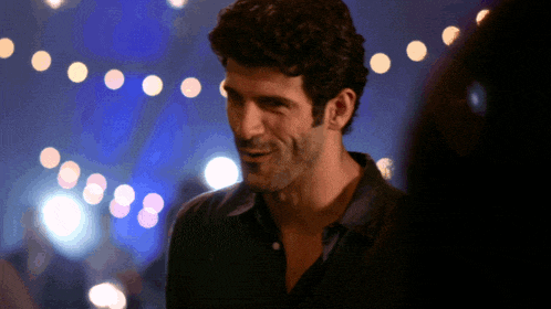 a man in a black shirt stands in front of a string of lights and smiles