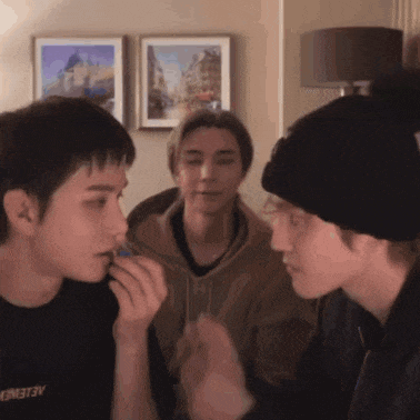 three young men are having a conversation in a living room and one of them is wearing a black shirt that says " aelene "
