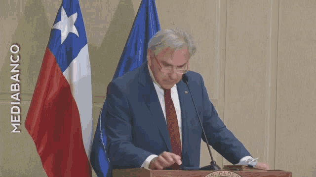 a man in a suit and tie stands at a podium in front of two flags and the words mediabanco on the wall behind him