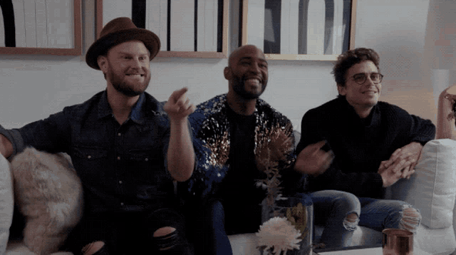 three men are sitting on a couch and one of them is wearing a hat