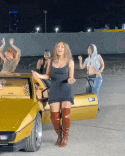 a group of women are dancing in front of a yellow sports car