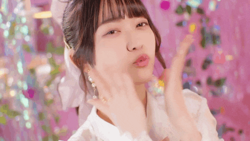 a girl in a white dress is blowing a kiss at the camera while standing in front of a pink background .