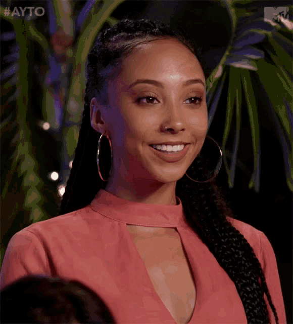 a woman wearing hoop earrings and a pink top smiles in front of a palm tree