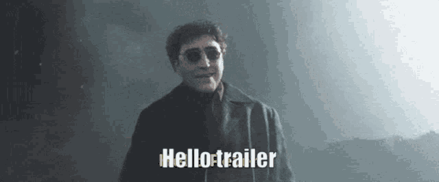 a man wearing sunglasses and a black coat is standing in a dark room and says hello trailer .