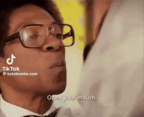 a man wearing glasses and a tie is talking to another man and says `` open your mouth '' .