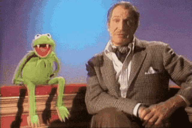 a man in a suit sits on a couch next to a kermit the frog puppet