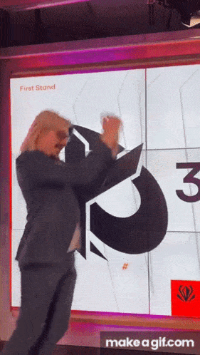 a woman in a suit is dancing in front of a large screen that says first stand