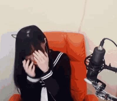 a girl in a school uniform is sitting in an orange chair in front of a microphone covering her face .